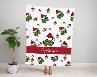 May include: White blanket featuring a repeating pattern of a green Grinch wearing a Santa hat, red bows, and bells. A red band with the name "Anderson" is across the bottom. The blanket is a festive holiday item.