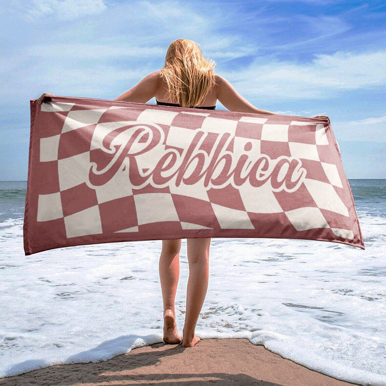 May include: A beach towel with a checkered pattern in burgundy and cream, held up on a beach. The towel features the name "Rebbica" in a cursive font.