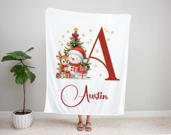 May include: White Christmas blanket featuring a snowman, reindeer, Christmas tree, and gifts. A large red letter "A" and the name "Austin" are also displayed. The blanket is held up by a person.