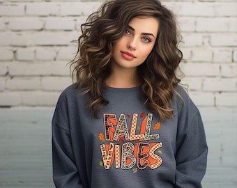 May include: A dark gray sweatshirt with the words "FALL VIBES" in a colorful, autumn-themed design. The text incorporates plaid, leopard print, and leaf accents. The sweatshirt is a casual fall fashion item.
