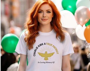 May include: A white t-shirt with the text "Charlie Kirk A True Patriot" and a golden dove design. The shirt also includes the phrase "In Loving Memory Of". The person in the image has red hair and is smiling. Balloons in green, white, and orange are in the background.