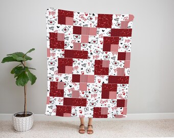May include: A white and red patchwork blanket with a Valentine's Day theme. The blanket features red squares with various patterns, including hearts, stripes, and floral designs. The words "love" and "xoxo" are also printed on the blanket.