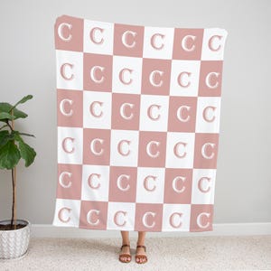 May include: A pink and white checkered throw blanket with the letter "C" repeated in a pattern.