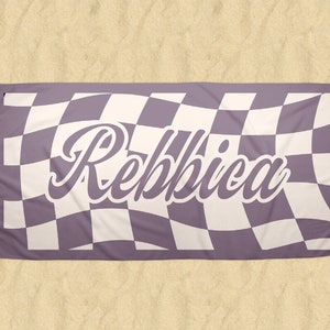 May include: A rectangular beach towel with a checkered pattern in lavender and white. The word "Rebbica" is written in a cursive font across the center of the towel. The towel is lying on a sandy surface.