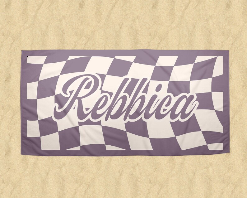 May include: A rectangular beach towel with a checkered pattern in lavender and white. The word "Rebbica" is written in a cursive font across the center of the towel. The towel is lying on a sandy surface.