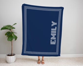 May include: A navy blue blanket with a white border and the name "EMILY" printed vertically in white. The blanket is rectangular and appears to be made of soft material. A potted plant is visible to the left.