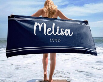 May include: A navy blue beach towel with white trim and the name "Melisa" in white script. The year "1990" is below the name. The towel is held up by a person on a sandy beach with the ocean in the background.