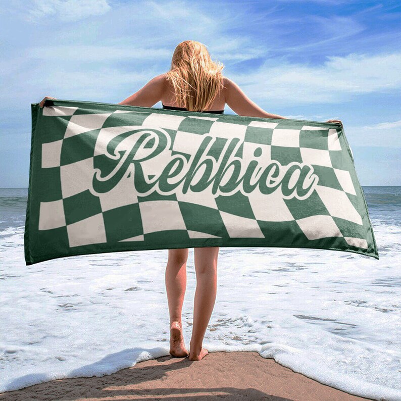 May include: A beach towel with a green and white checkered pattern, held up by a person on a sandy beach. The towel features the name "Rebbica" in a cursive font across the center. The ocean and sky are visible in the background.