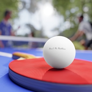 Puede incluir: Una pelota de ping pong blanca con la inscripción "Mrs & Mr Matthew" descansa sobre una paleta roja. La paleta está sobre una mesa azul, con una red y figuras borrosas en el fondo.