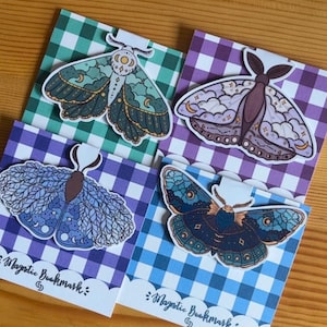 May include: Four magnetic bookmarks featuring illustrated moths with celestial patterns. Each moth is attached to a square card with a gingham check pattern in green, purple, or blue. The cards feature the text Magnetic Bookmark at the bottom.
