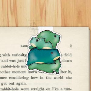 May include: A magnetic bookmark featuring a stack of three illustrated frogs in shades of light green, teal, and dark green. The frogs have black eyes and cream-coloured bellies. A small logo with the text The Crowned Collective is visible.