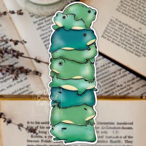 May include: A sticker depicting a stack of seven cartoon frogs in shades of green and teal. The frogs are stacked, creating a whimsical design. The sticker has a white border and the text "The Crowed Collective" is visible.