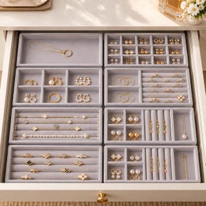 May include: A set of light gray velvet jewelry organizer trays arranged inside a drawer. The trays feature various compartments, including slots for rings, earrings, and necklaces, filled with gold-toned jewelry pieces like chains, studs, and hoops.