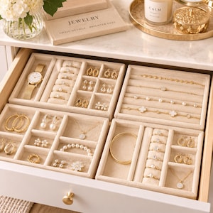 May include: A beige velvet jewelry organizer tray set inside a drawer, containing gold-toned watches, rings, necklaces, bracelets, and earrings. The compartments are lined with soft fabric, neatly displaying various pieces of jewelry in organized sections.