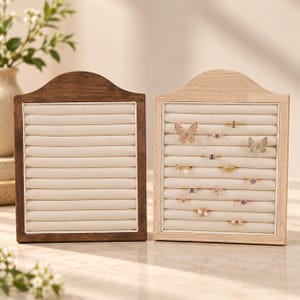 May include: Two rectangular ring display holders with arched tops, one in dark brown wood and one in light natural wood. Each features a cream-colored velvet insert with horizontal slots for organizing rings, with several rings displayed.