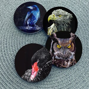 May include: A set of four circular coasters featuring detailed illustrations of birds. One coaster shows a blue-toned crow, another a bald eagle, a third a great horned owl with yellow eyes, and the fourth a black swan with a bright red beak.