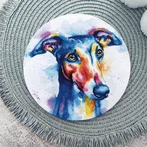 May include: A circular print featuring a watercolour painting of a dog with large brown eyes. The artwork uses vibrant splashes of blue, yellow, orange, and pink tones to depict the canine's face, set against a white background on the circular surface.