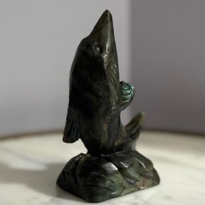 Puede incluir: Escultura de piedra tallada de color verde oscuro de un pez saltando hacia arriba. La escultura tiene una superficie texturizada y est&aacute; montada sobre una base. El pez tiene un ojo y aletas detallados. La escultura mide unos 13 cm de altura.