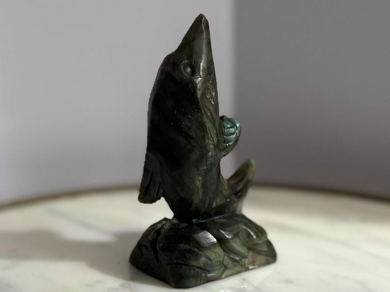 Puede incluir: Escultura de piedra tallada de color verde oscuro de un pez saltando hacia arriba. La escultura tiene una superficie texturizada y est&aacute; montada sobre una base. El pez tiene un ojo y aletas detallados. La escultura mide unos 13 cm de altura.