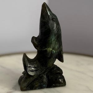 Puede incluir: Una escultura de delf&iacute;n de piedra tallada en tonos verdes y negros. El delf&iacute;n est&aacute; representado saltando hacia arriba, con la boca abierta y las aletas extendidas. La escultura es detallada y se asienta sobre una superficie similar al m&aacute;rmol.