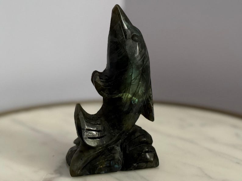 Puede incluir: Una escultura de delf&iacute;n de piedra tallada en tonos verdes y negros. El delf&iacute;n est&aacute; representado saltando hacia arriba, con la boca abierta y las aletas extendidas. La escultura es detallada y se asienta sobre una superficie similar al m&aacute;rmol.