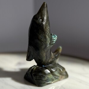 Puede incluir: Una escultura de delf&iacute;n de piedra tallada en tonos grises y verdes. El delf&iacute;n est&aacute; representado saltando de una base, sosteniendo una peque&ntilde;a bola. La escultura tiene un acabado pulido y est&aacute; sobre un fondo neutro.