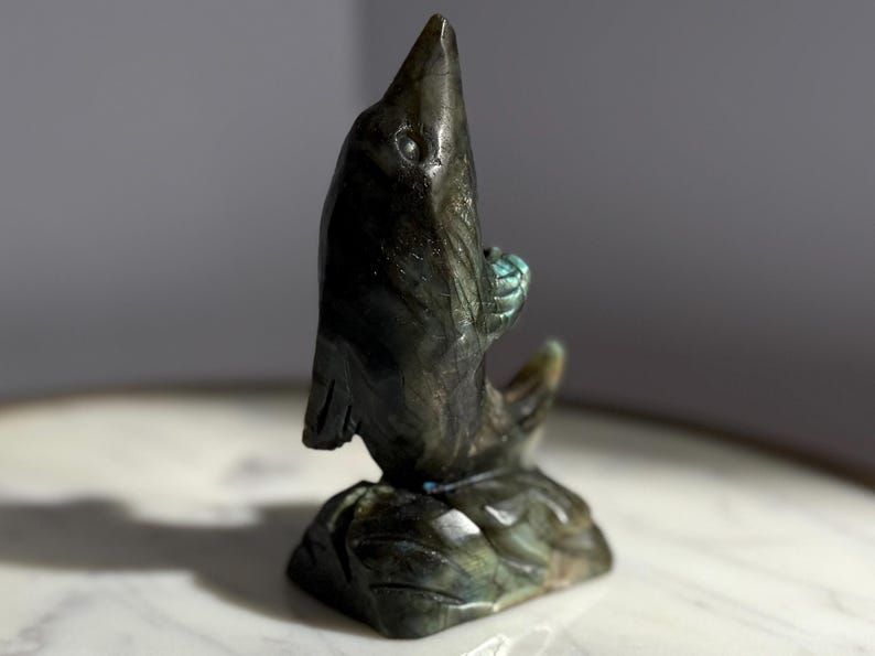 Puede incluir: Una escultura de delf&iacute;n de piedra tallada en tonos grises y verdes. El delf&iacute;n est&aacute; representado saltando de una base, sosteniendo una peque&ntilde;a bola. La escultura tiene un acabado pulido y est&aacute; sobre un fondo neutro.