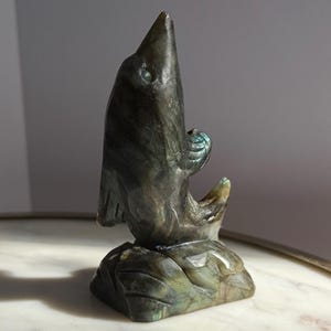 Puede incluir: Una escultura de piedra tallada de un p&aacute;jaro con un pico largo y una forma estilizada. La escultura es principalmente de color gris oscuro con toques de azul y verde, y se asienta sobre una base cuadrada. El p&aacute;jaro mira hacia arriba.