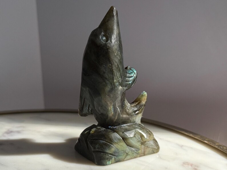 Puede incluir: Una escultura de piedra tallada de un p&aacute;jaro con un pico largo y una forma estilizada. La escultura es principalmente de color gris oscuro con toques de azul y verde, y se asienta sobre una base cuadrada. El p&aacute;jaro mira hacia arriba.