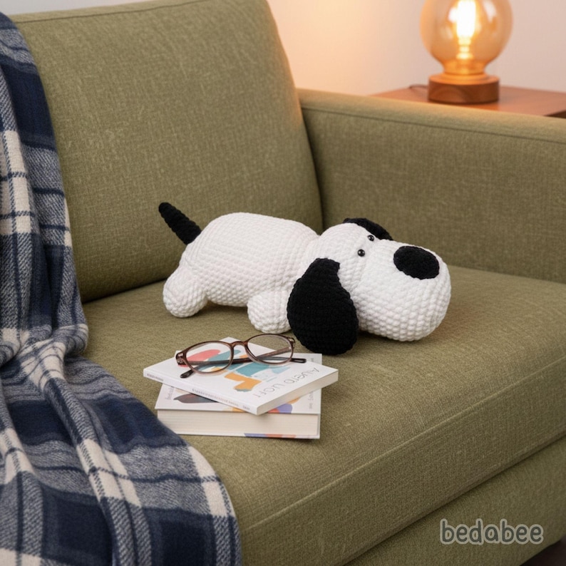 May include: A white and black crochet dog plush toy with black ears and a black tail is lying on a green sofa. The dog is resting on a stack of books and a pair of glasses. A blue and white plaid blanket is draped over the arm of the sofa.