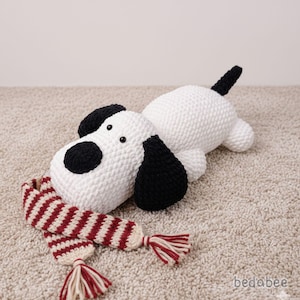 May include: A handmade, white and black crocheted dog toy with a red and cream striped scarf. The dog has black ears and a black spot over one eye. The scarf has tassels on each end. The dog is lying down.