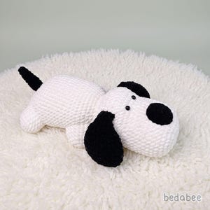 May include: A handmade white and black crochet dog toy. The dog has black ears, a black tail, and a black spot on its face. The toy is lying on a fluffy white surface. The brand name "bedabee" is visible in the bottom right corner.