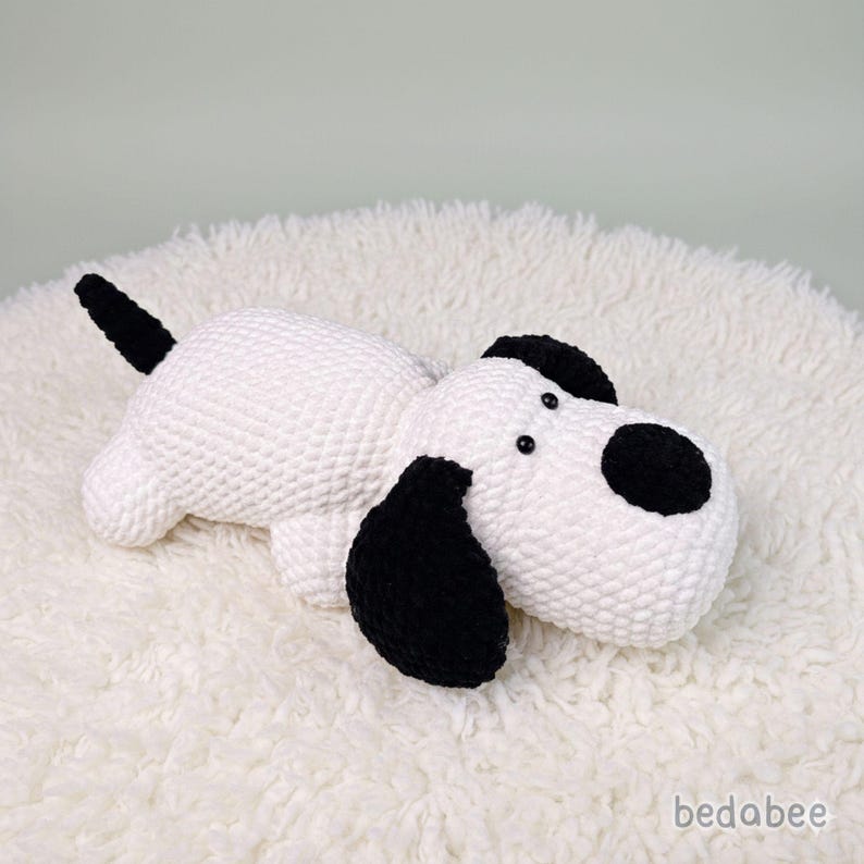 May include: A handmade white and black crochet dog toy. The dog has black ears, a black tail, and a black spot on its face. The toy is lying on a fluffy white surface. The brand name "bedabee" is visible in the bottom right corner.