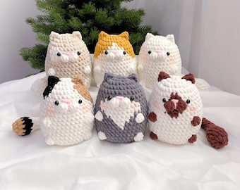 May include: Six crocheted cat plushies in various colors, including white, brown, gray, beige, and orange. The plushies are arranged in a semi-circle on a white surface.