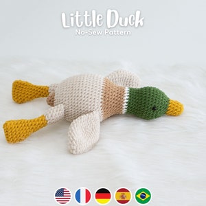 May include: A crocheted stuffed duck toy lying on a white fluffy surface. The duck is beige, brown, green, and yellow. The duck has a long beak and small black eyes. The text "Little Duck No-Sew Pattern" is above the duck.