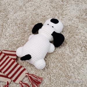 May include: A white and black knitted dog toy with black ears and spots. A red and white striped scarf with tassels is next to the dog. The dog is lying on a beige carpet.