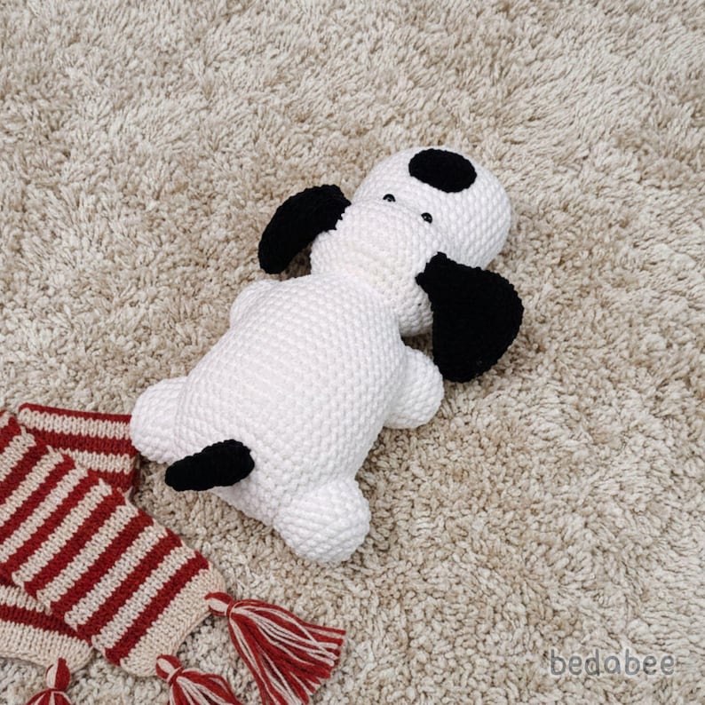 May include: A white and black knitted dog toy with black ears and spots. A red and white striped scarf with tassels is next to the dog. The dog is lying on a beige carpet.