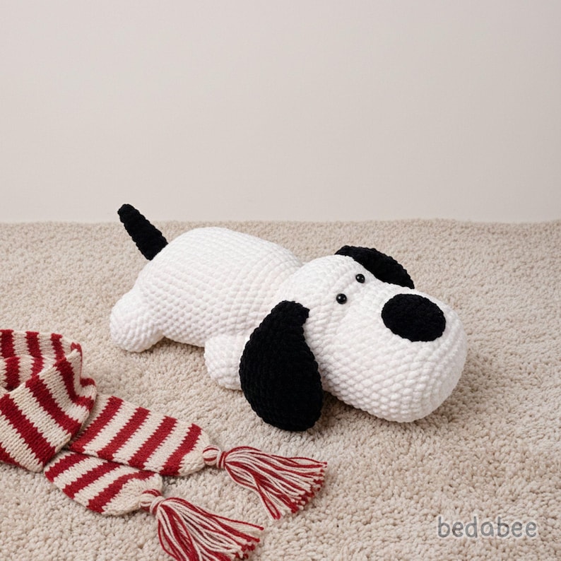 May include: A white and black plush dog toy with black ears and a black tail. The dog is lying on a beige carpet next to a red and white striped scarf with tassels. The dog has black button eyes.