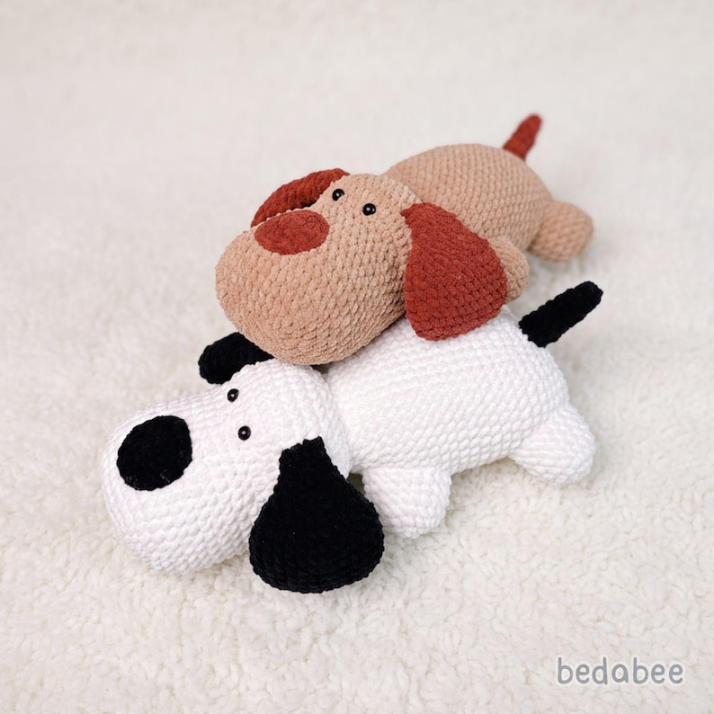 May include: Two handmade crochet dog toys. One is tan with red-brown ears and nose, the other is white with black spots. The toys are lying on a white, textured surface. The brand name "bedabee" is in the bottom right corner.