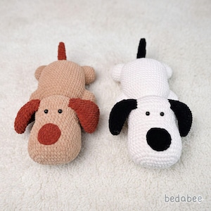 May include: Two handmade crochet dog toys. One is tan with rust-colored ears and nose, the other is white with black ears and nose. Both have small black eyes and a tail. The toys are lying on a white, textured surface.