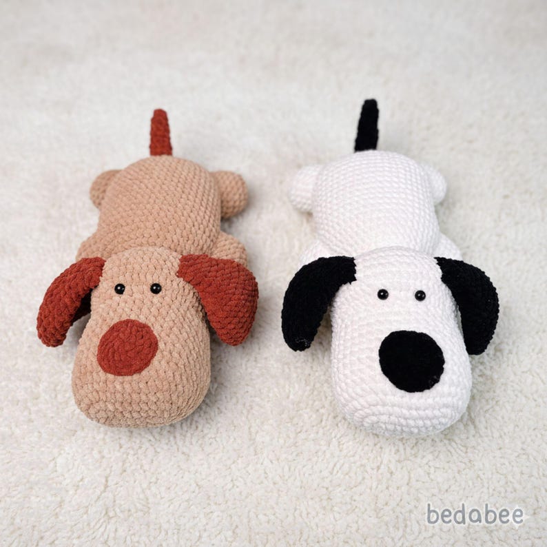 May include: Two handmade crochet dog toys. One is tan with rust-colored ears and nose, the other is white with black ears and nose. Both have small black eyes and a tail. The toys are lying on a white, textured surface.