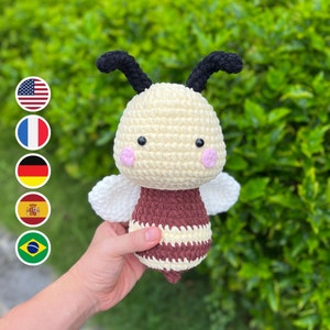 May include: A crocheted bee toy with a white body, brown and white striped abdomen, and black antennae. The bee has pink cheeks and black eyes.