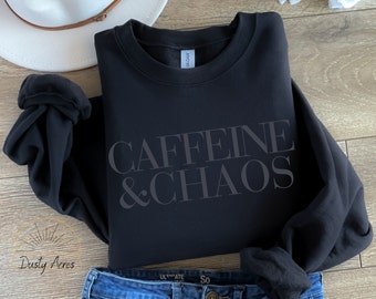 May include: Black crewneck sweatshirt with the text "CAFFEINE & CHAOS" in a dark gray font. The sweatshirt is folded, with a white hat and blue jeans in the background. The brand name "Dusty Acres" is in the lower left corner.