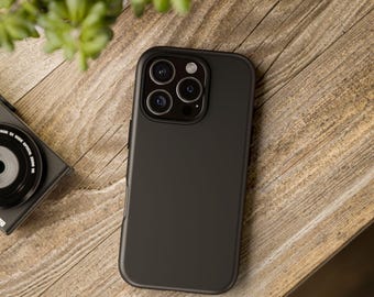 以下が含まれることがあります： マット仕上げの黒いスマートフォンケース。ケースには、複数のレンズを備えた正方形のカメラモジュールがあります。携帯電話ケースは木製の表面に置かれています。小さなカメラと植物も表示されます。