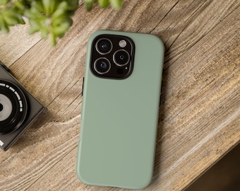 May include: A sage green phone case with a black camera module rests on a wooden surface. The phone case has a smooth, matte finish. A black camera and a green plant are also visible.