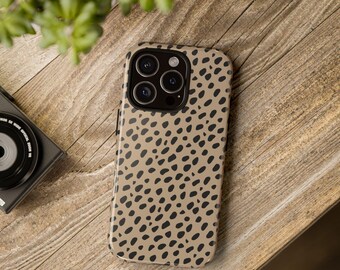 Könnte beinhalten: Eine beige Handyhülle mit einem schwarzen Fleckenmuster. Die Hülle liegt auf einer Holzoberfläche, mit einer Kamera und Grünzeug im Hintergrund. Die Handyhülle hat ein schützendes Design.