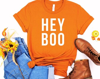May include: Orange t-shirt with the text "HEY BOO" in white letters. The shirt is laid flat on a white background with blue jeans, brown boots, and fall decor.
