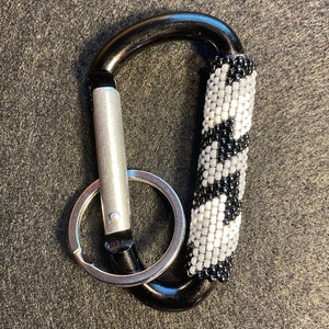 Könnte beinhalten: Schwarzer Karabinerhaken mit silbernem Verschluss und Schlüsselring. Der Griff ist mit einem Perlenmuster in Schwarz und Weiß verziert. Ein funktionelles Accessoire.