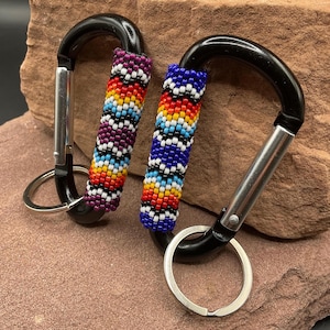 May include: Two black carabiner keychains with beaded accents. One keychain has a purple, red, orange, yellow, and white beaded design. The other has a blue, red, orange, yellow, and white beaded design. Both have silver key rings.