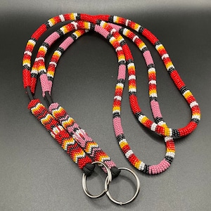 Peut inclure: Lanière perlée faite à la main en rouge, rose, jaune, noir et blanc. La lanière présente un motif répétitif de perles colorées et comprend deux anneaux métalliques argentés pour attacher des clés ou un badge d'identification.
